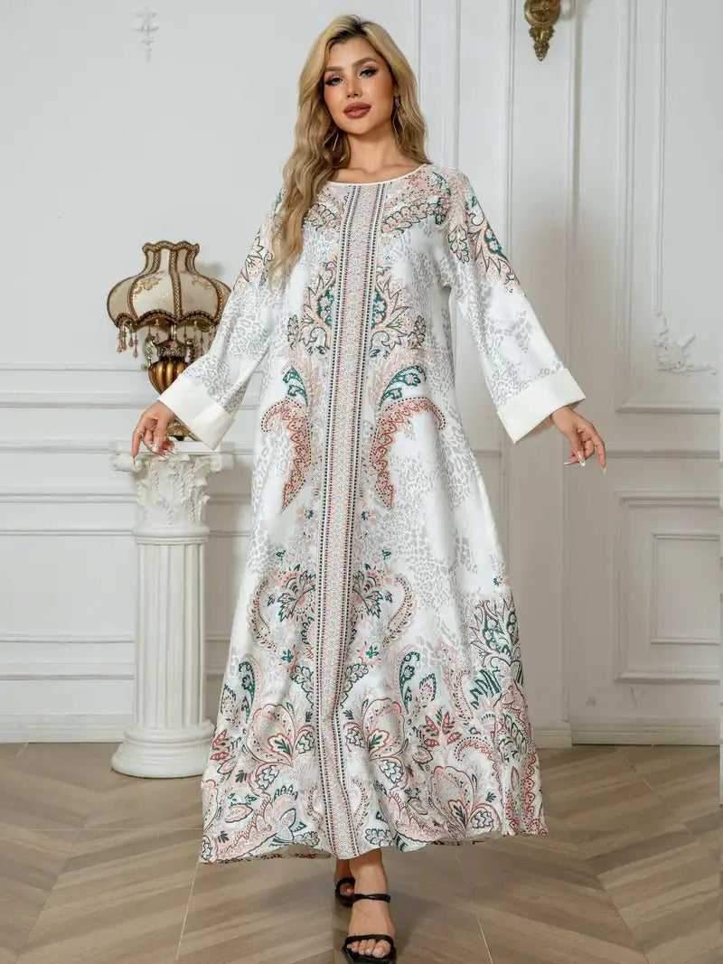 A woman modeling a long white kaftan dress with intricate multicolor paisley and floral embroidery, standing in a classic white interior room with wood floors.