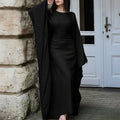 Woman wearing a long black coat over a black dress against a stone wall and white door background