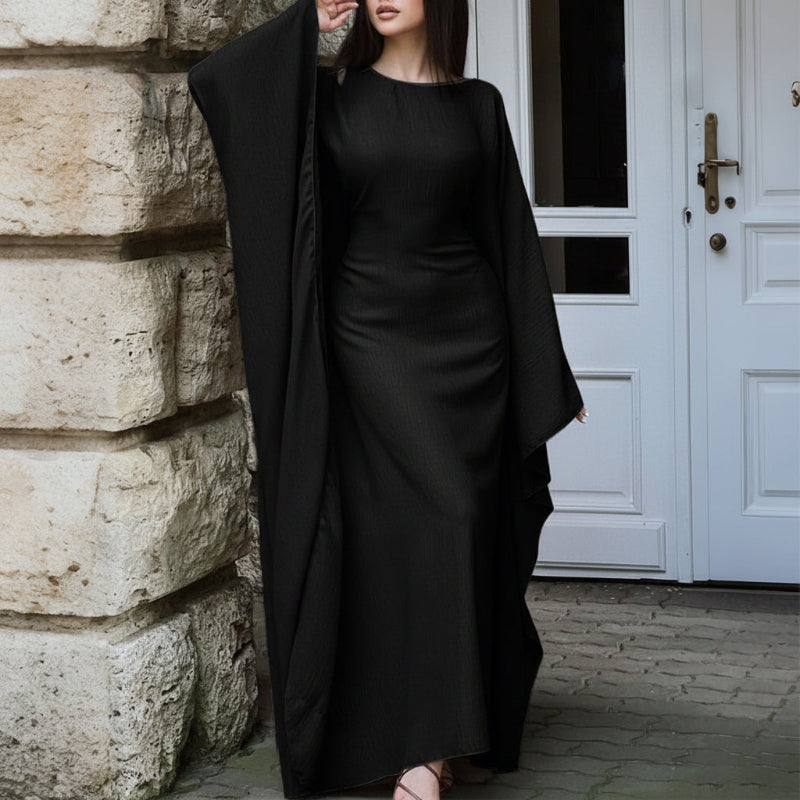 Woman wearing a long black coat over a black dress against a stone wall and white door background