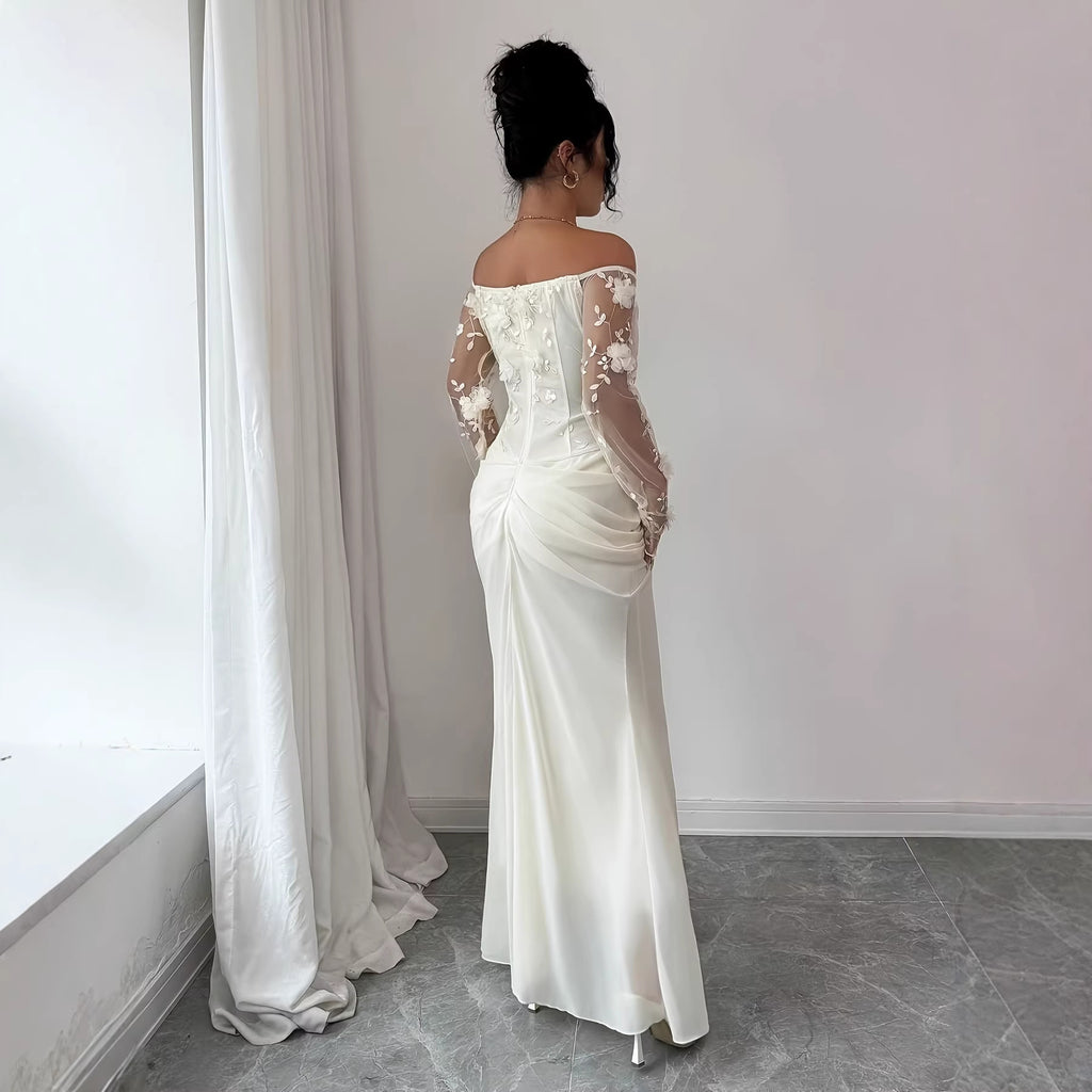 A woman stands with her back to the camera wearing an off-the-shoulder ivory wedding dress with sheer floral lace sleeves and a ruched satin skirt, in a minimalist room with white walls and gray floor tiles.