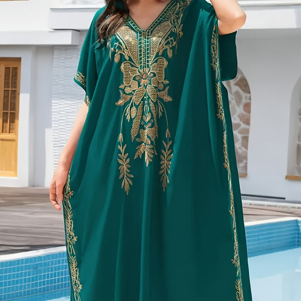 A woman wearing a flowing teal green kaftan dress with ornate gold embroidery, standing by a swimming pool in front of a white building.