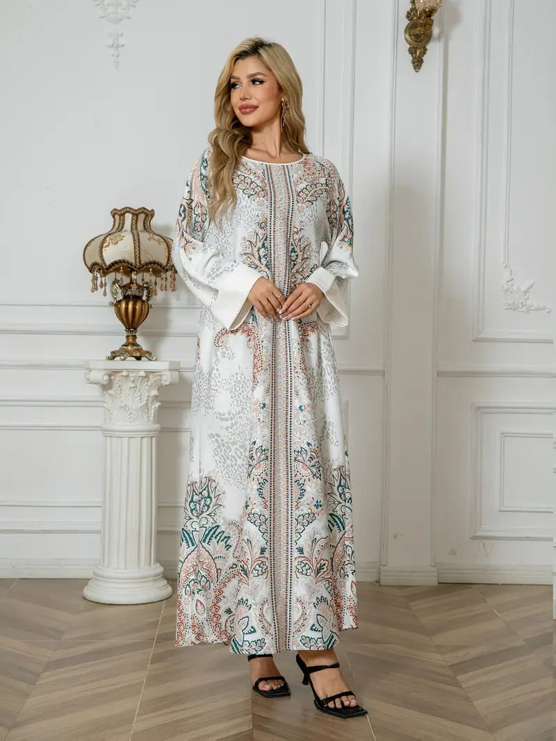 A woman models a long white kaftan dress with intricate multicolor floral embroidery in blue, pink, and green, standing in a classic white interior room with a wooden herringbone floor.