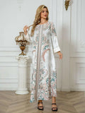 A woman models a long white caftan dress with a detailed multicolor paisley and floral pattern in pink, green, and blue, styled with black strappy sandals in an elegant white room.