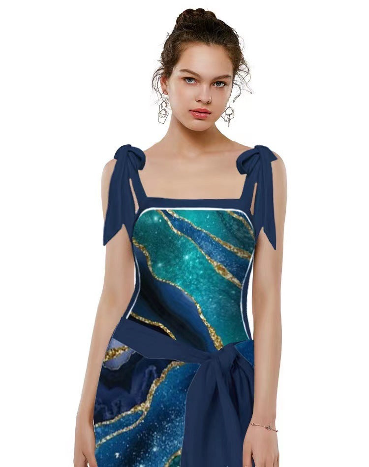 A model wearing a sleeveless navy blue dress with a teal and gold agate-like patterned bodice and skirt, featuring tie details at the shoulders and a large waist bow, set against a plain white background.