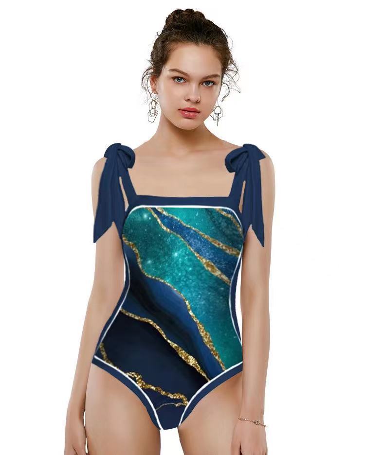 A model wearing a one-piece swimsuit with a navy blue base, teal and green marble-like patterns, and metallic gold veining, featuring navy shoulder ties and white piping.