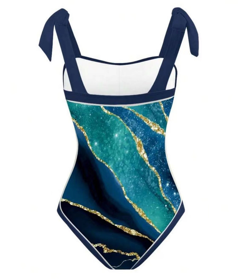 A one-piece swimsuit with a navy blue and teal marble pattern accented by gold veins, featuring a white inner lining and navy blue tie shoulder straps, shown on a plain white background.