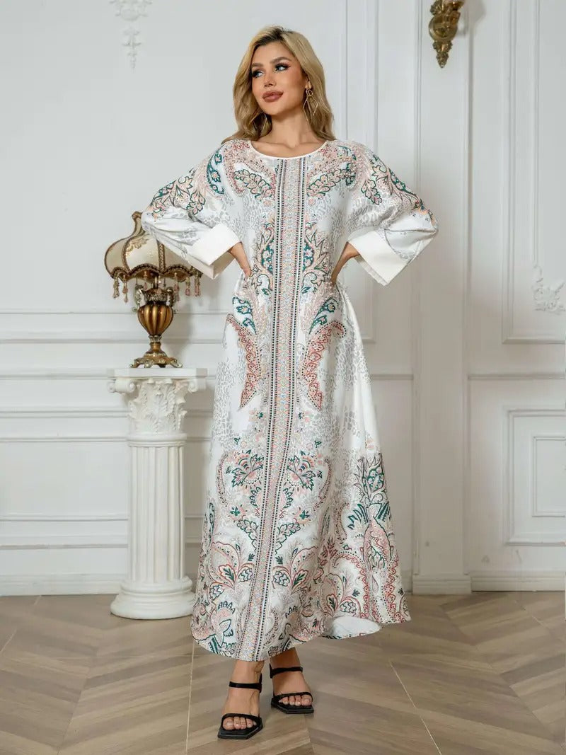 A woman models a long white kaftan dress with intricate multicolor paisley embroidery in pink, teal, and gold, standing in a classic white interior room with a wooden herringbone floor.