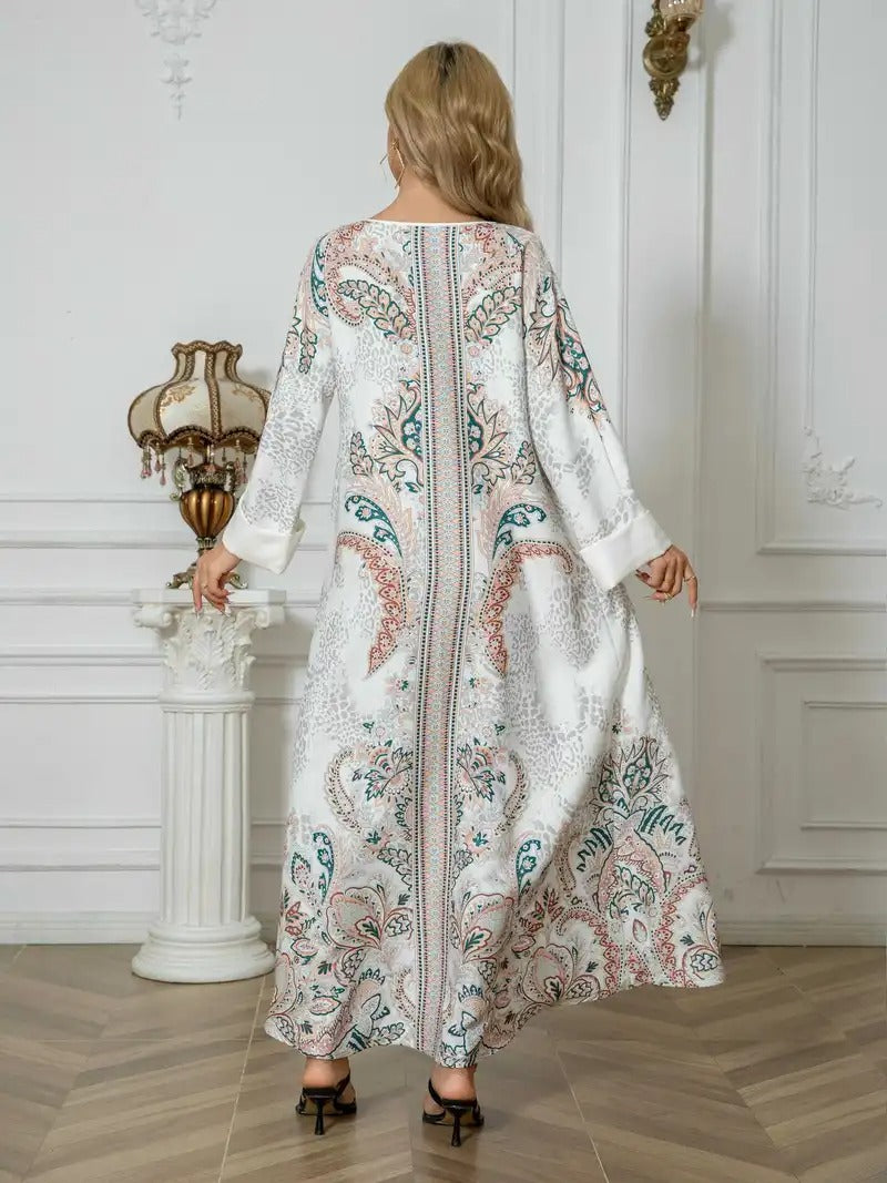 A woman viewed from behind wearing a long white kaftan dress with intricate teal and dusty pink paisley patterns, standing in a classic white interior room with wooden floors.