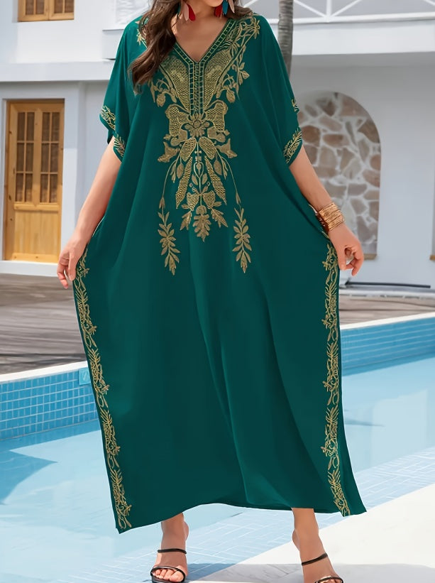 A woman wearing a flowing dark green kaftan dress with gold embroidery, standing by a swimming pool in front of a white building.