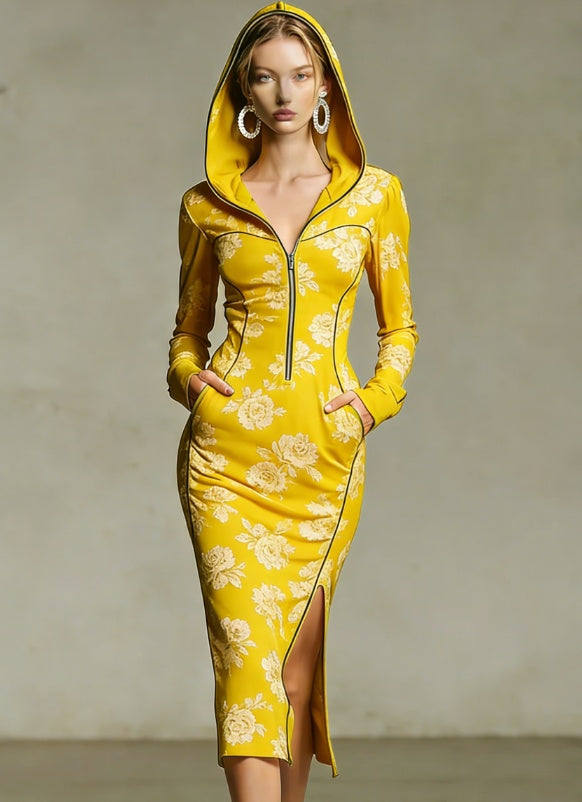 A woman models a bright yellow hooded long-sleeve dress with a white floral pattern, front zipper, and thigh-high slit, set against a neutral gray studio background.